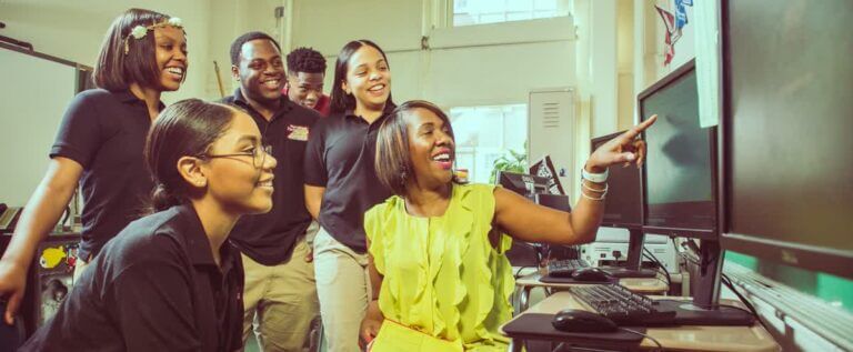A CBI director of college counsleling sits at a computer sorrounded by students; she points at something on the monitor and they look on