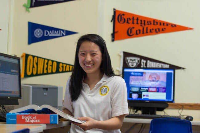 A student stands in the middle of a college counseling office. She has a book open in front of her and she is looking up, smiling at the camera.