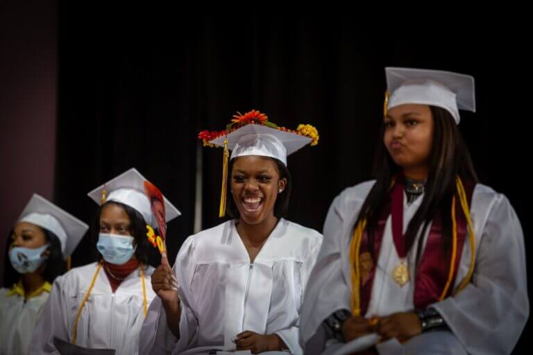 There are four graduates wearing white gowns. One has flowers on her cap. They are all smiling and looking away from the camera.