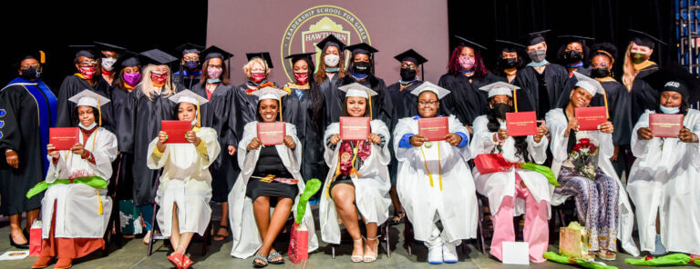 The first graduates of Hawthorne sit on a stage, holding up their diplomas. Teachers and school staff are standing and smiling behind the graduates.