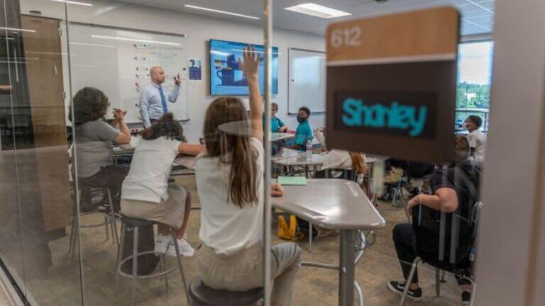 A grtoup of students are inside a classroom with a teacher standing at the front of the room. The students are raising their hands and appear to be engaged in their learning.