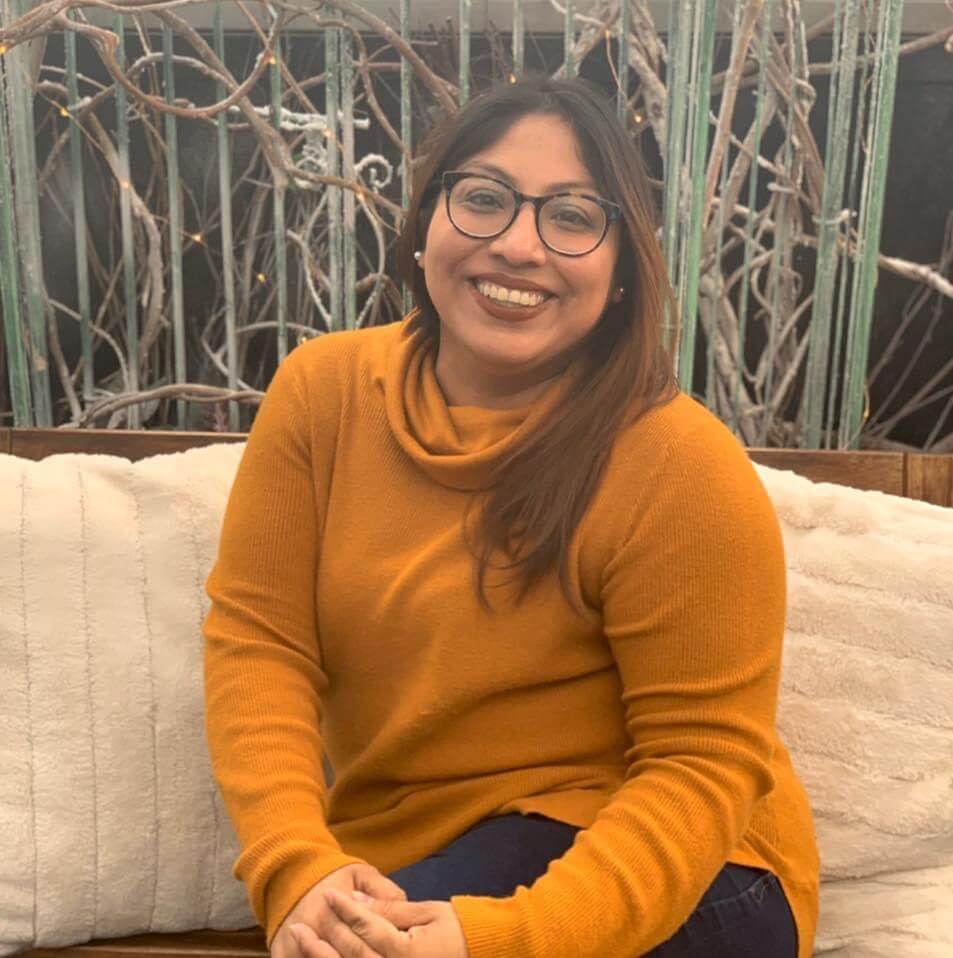 Alendi is sitting on a sofa with a lovely orange sweater and is warmly smiling at the camera.