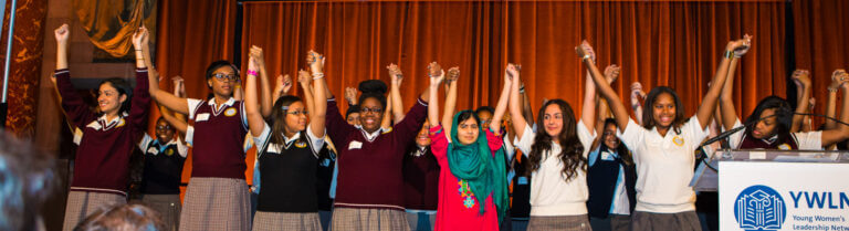 Malala stands in the center of a line of TYWLS students. They are holding hands with their arms triumphantly raised in the air