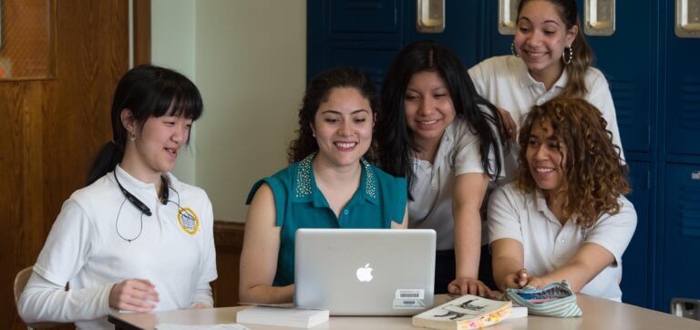 A teacher sits at a table with an Apple computer. Four TYWLS students are looking on and smiling.