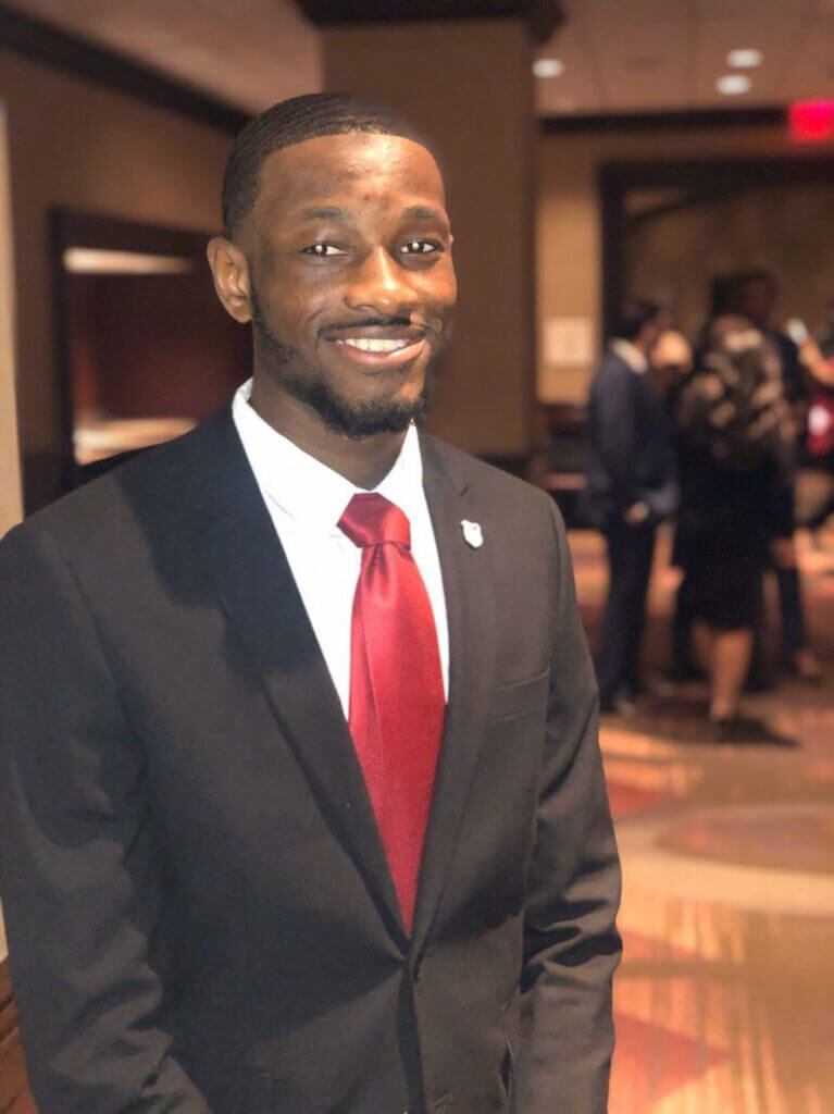 Basheeru Camara in a suit and a red tie. He is looking at the camera smiling. 
