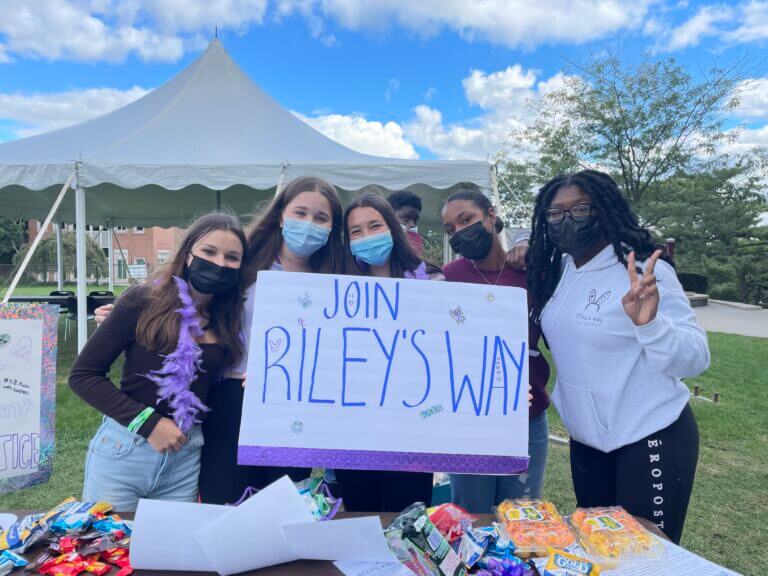 Students from TYWLS Bronx are outside at an event. They're holding up a sign that says "Join Riley's Way."