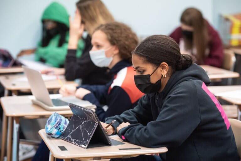 Students wearing masks are sitting at their desks, working on laptops and tablets.