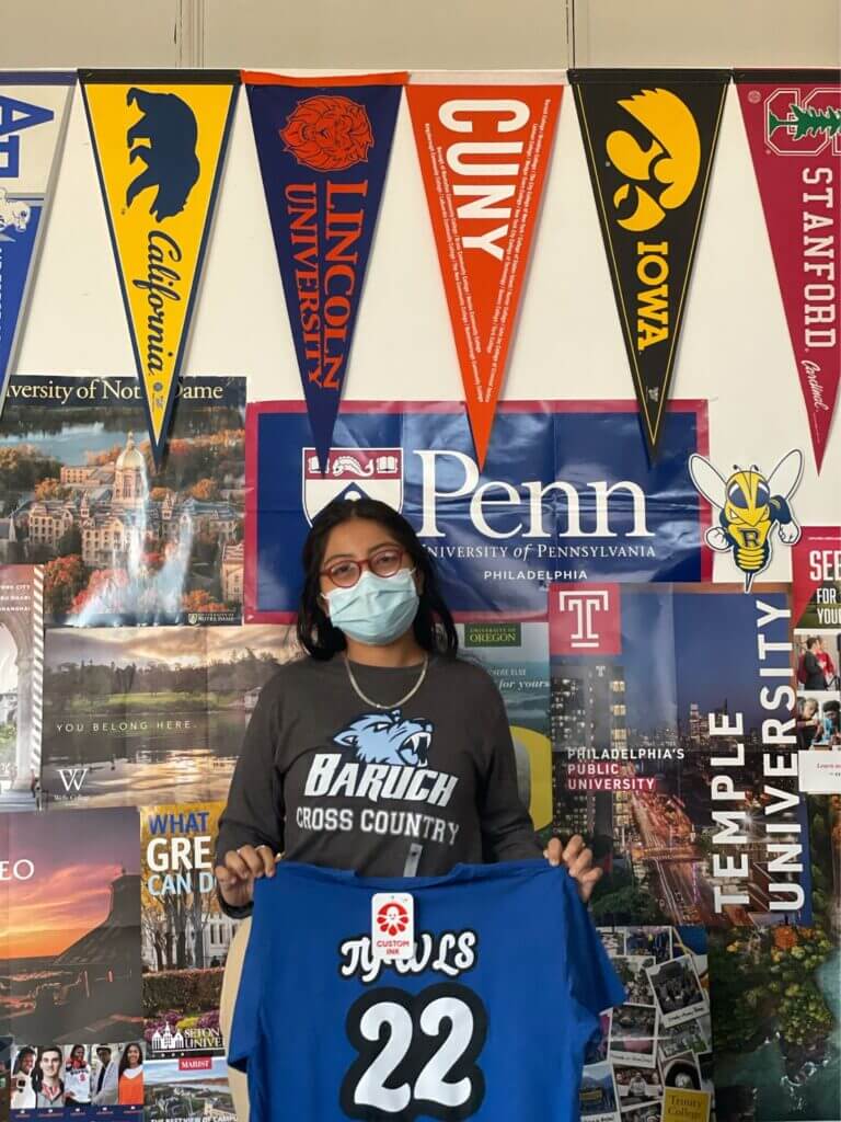 Jessica is standing in the college office at TYWLS Brooklyn. She is wearing a mask and holding a blue shirt that reads TYWLS '22.