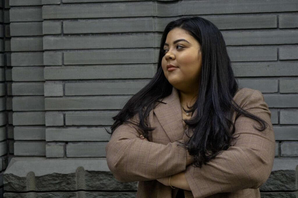 Priscilla is standing in front of a gray brick wall and looking off to the side. She has her arms folded and looks very powerful and confident. 
