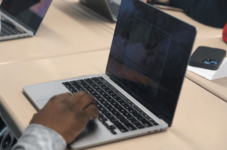 A Macbook sits on a table. There is a students hand touching the space bar.