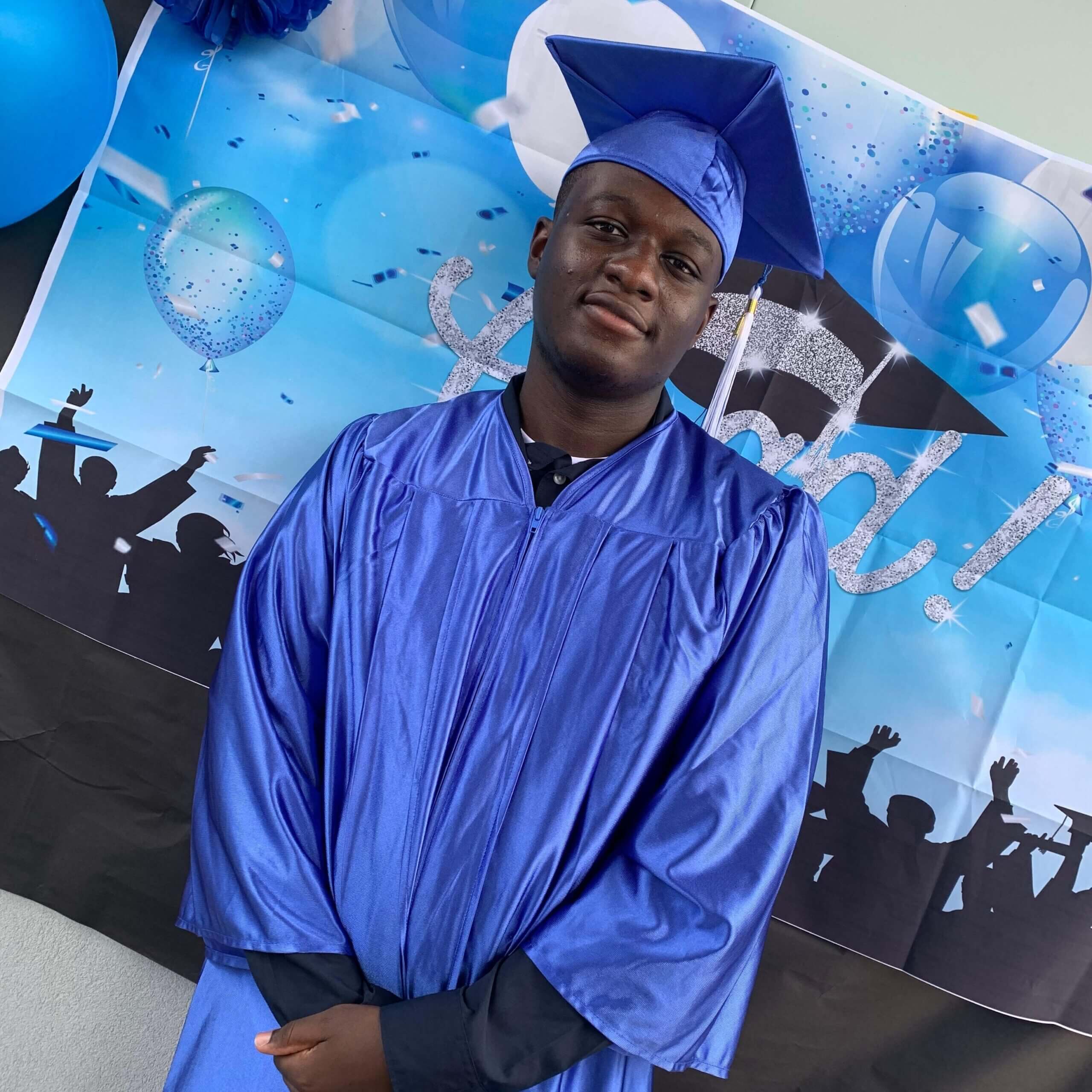 Kelvin is facing the camera, wearing a blue graduation cap and gown. In the background is a blue and silver graduation banner.