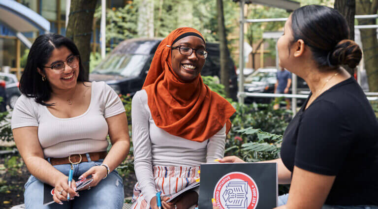 Three students sit on a bench and are laughing. They are sitting outside with a college building behind them.