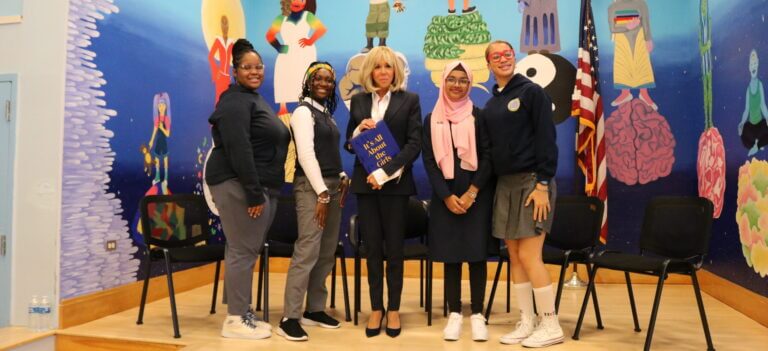 First Lady Macron stands in the middle of the four TYWLS students who moderated the panel discussion. Macron is holding a copy of "Its all about the girls"