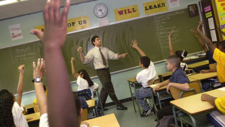 A teacher stands at the front of a classroom and several students have their hands raised.