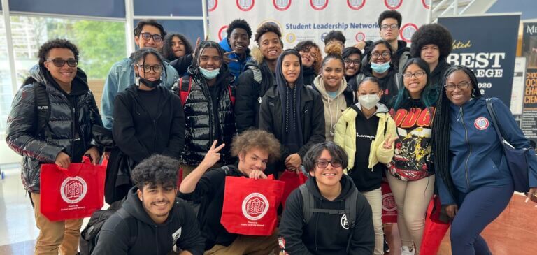 A group of seniors take a fun group picture with their director of college counseling. They are holding red CBI tote bags.