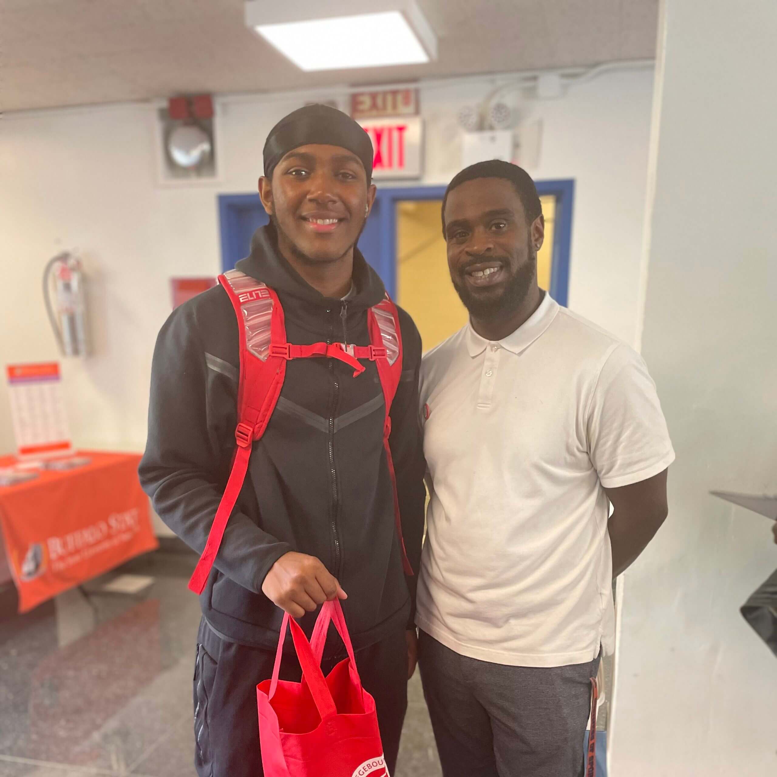 Tavonn stands next to his DCC, Jeffrey, for a photo. He is holding a red tote and wearing a red backpack. 