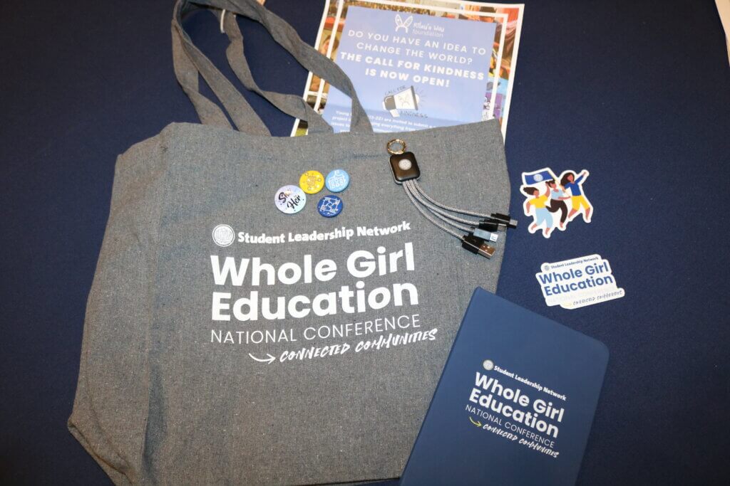 Branded items from the Whole Girl Education National Conference including a tote bag, notebook, pins, stickers, pen, etc.