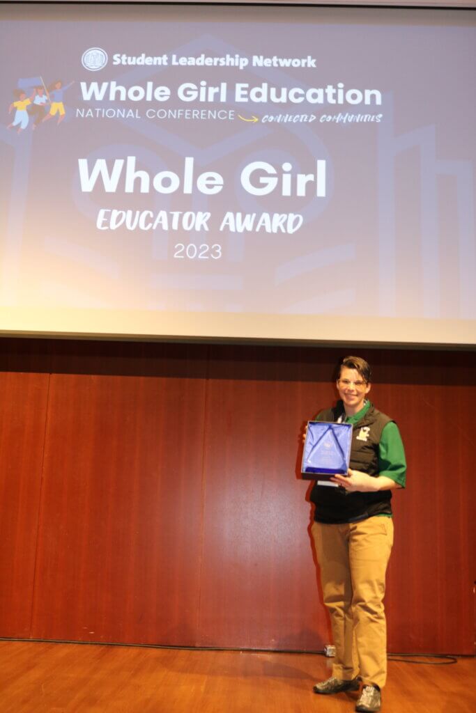 Jillian Jardas on stage holding an award with "Whole Girl Educator Award" on the screen behind her