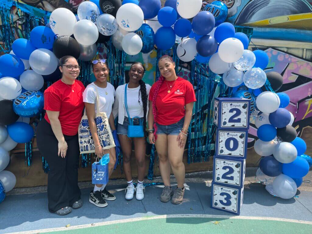 Joy poses with her friends under a balloon arch and a sign that reads 2023