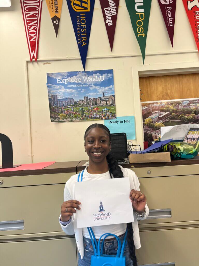 Joy stands under a streamer made up of various college pennants holding a sign that reads "Howard University"