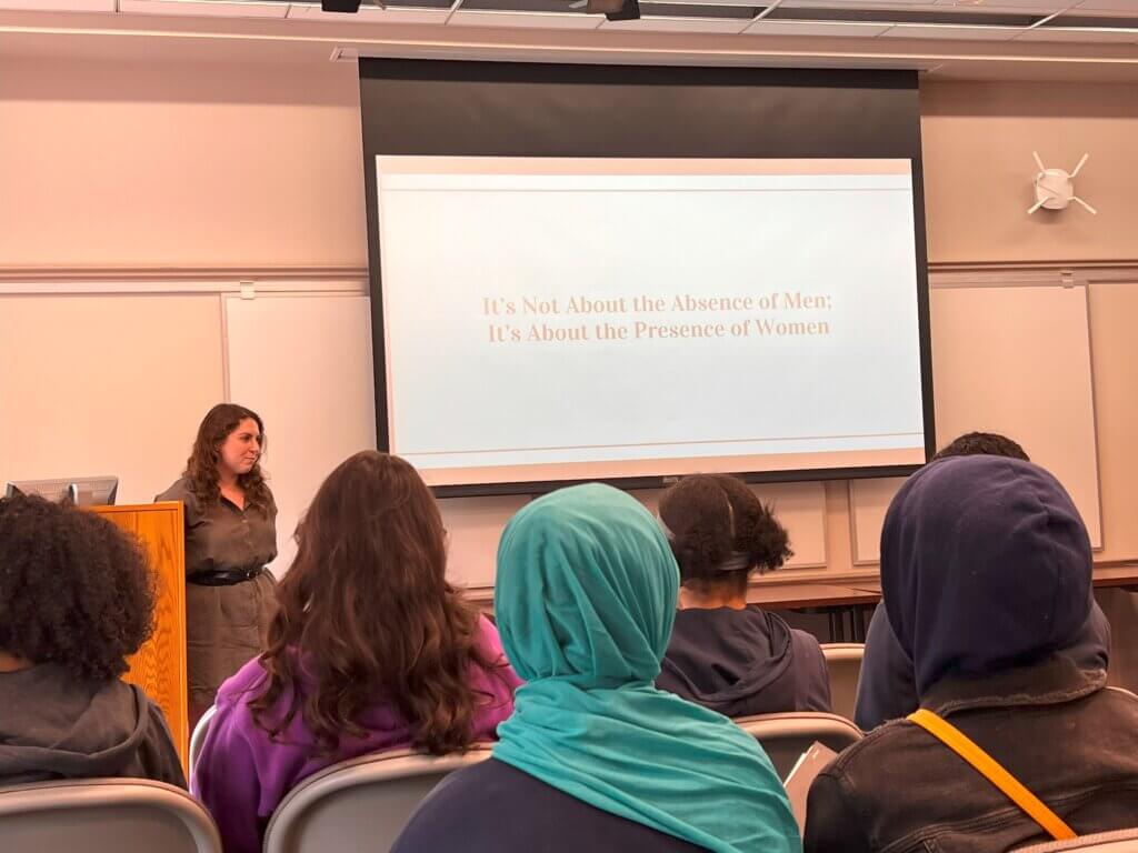 The words "It's not about the absence of men, it's about the presence of women" on a projection screen in a roomful of TYWLS students