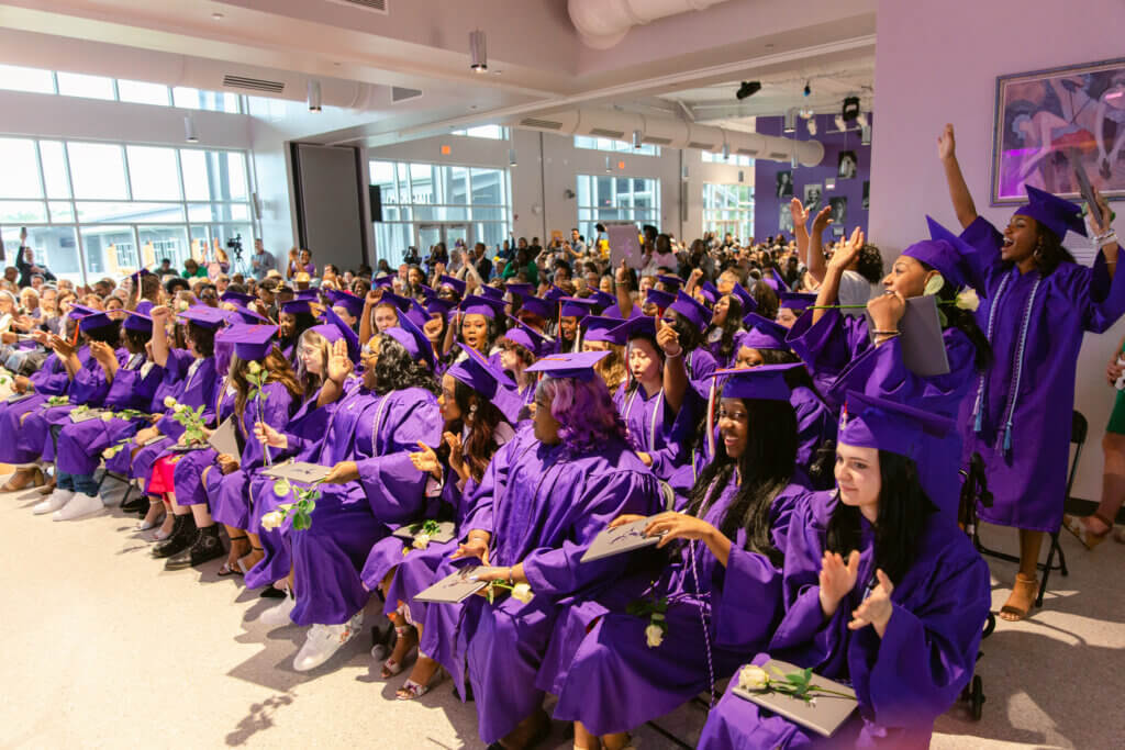 Students in purple graduation caps and gowns are seated in rows