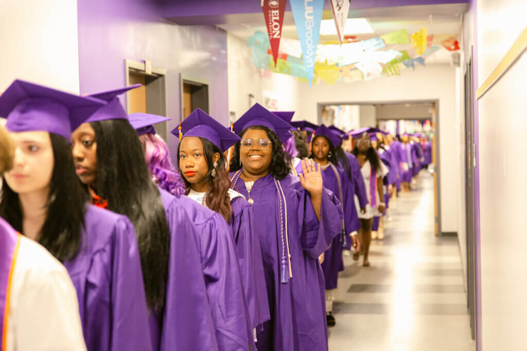 Girls in purple graduation caps and gowns line up in hallway