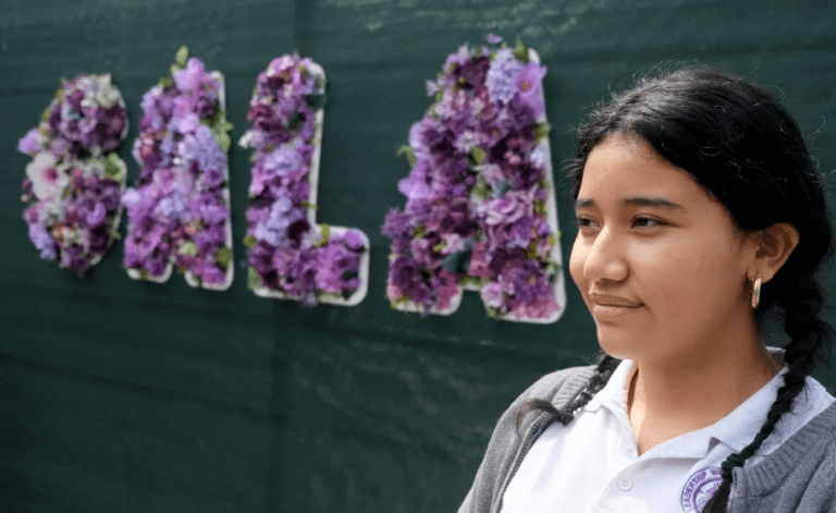 Student stands in front of school sign "GALA" which is covered by purple flowers