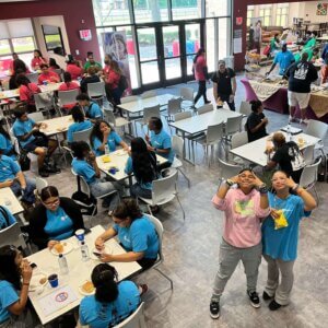 Groups of students in colorful t-shirts eat lunch, with two girls facing the camera on lower right.