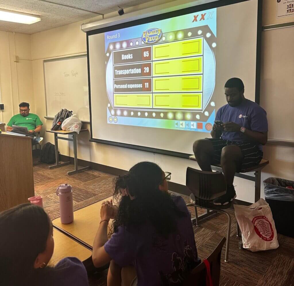 Two students and two counselors are inside a classroom, looking at a screen with trivia questions about college preparation.