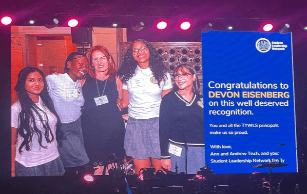 Large screen on a stage with photo of Principal Devon Eisenberg and her students, with congratulatory text on the right.