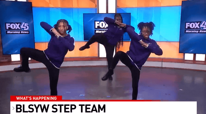 Three students from the Baltimore Leadership School for Young Women's step team perform on a television stage.