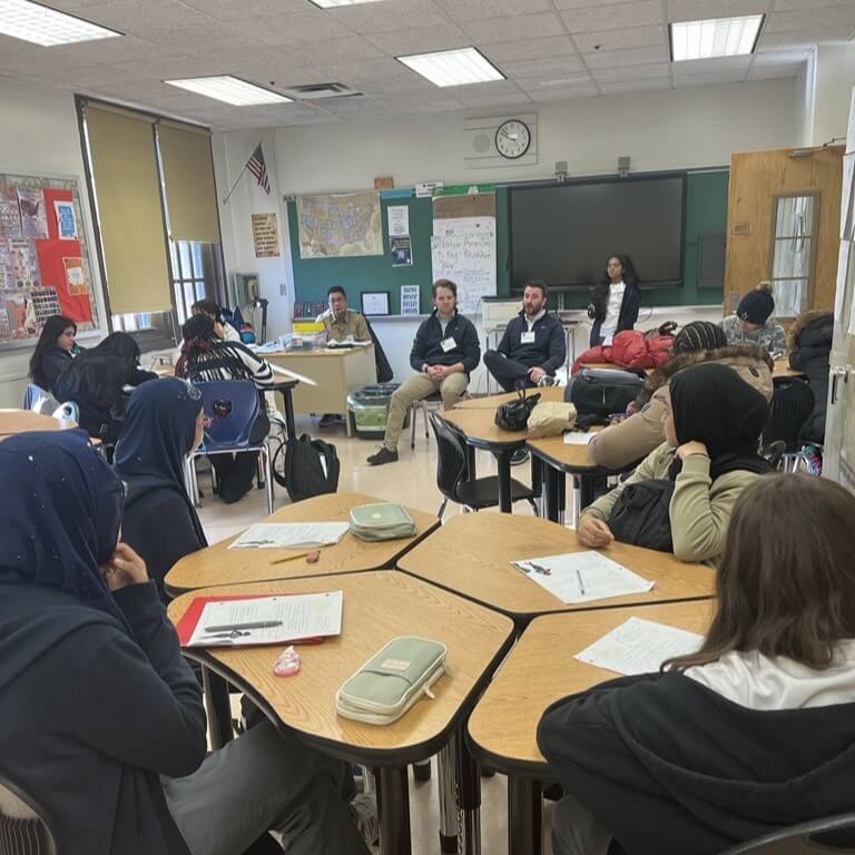 Two adult volunteers sit in front of classroom, with students facing them.