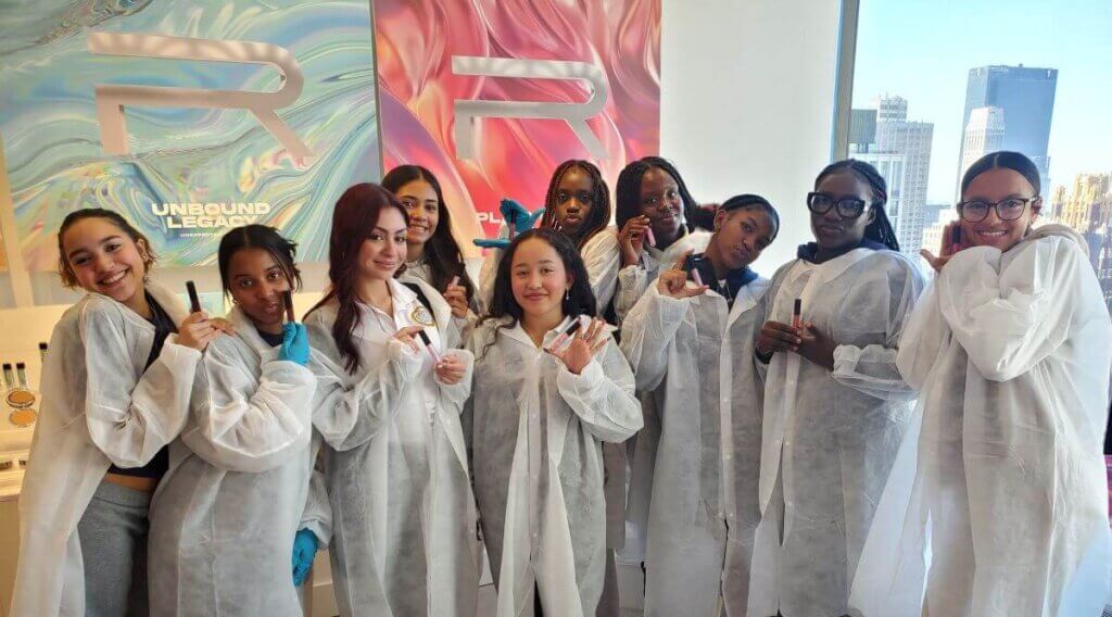 Ten young women wearing lab coats hold up the lip glosses they created.