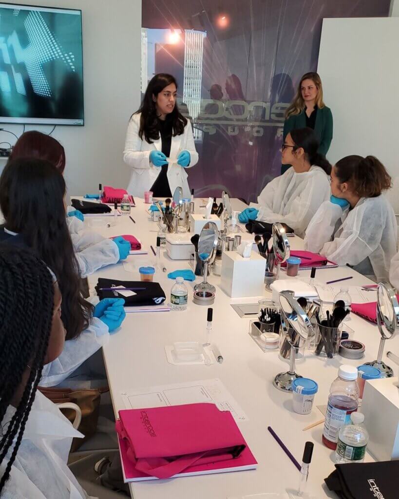 Female scientist and group of students, all in lab coats and gloves, measure out ingredients to create lipgloss.