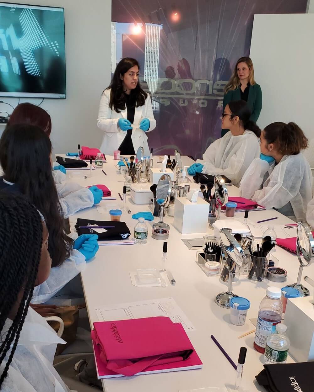 Female scientist and group of students, all in lab coats and gloves, measure out ingredients to create lipgloss.