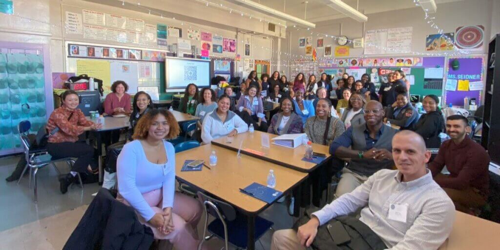 Over 30 adults and students in a classroom, some seated and some standing, facing camera for photo.