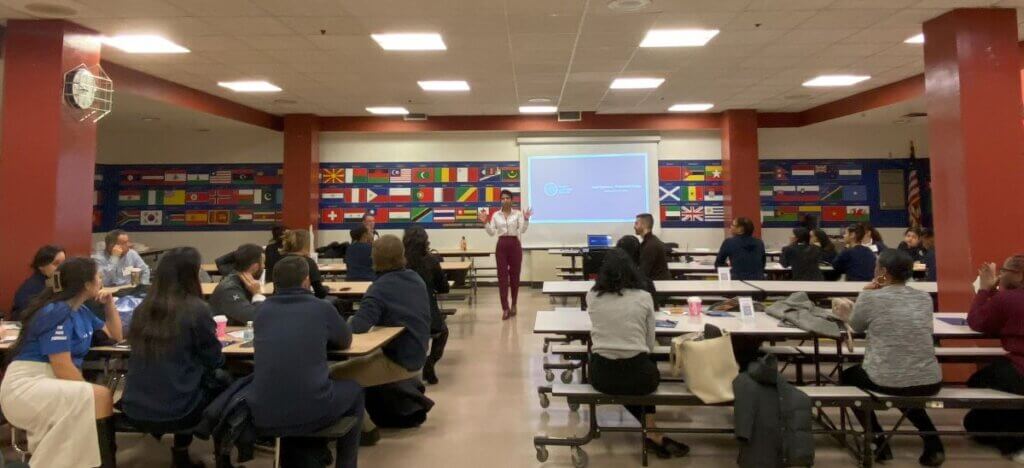 Principal stands in front of cafeteria for a speech, with over 20 adults sitting at tables.