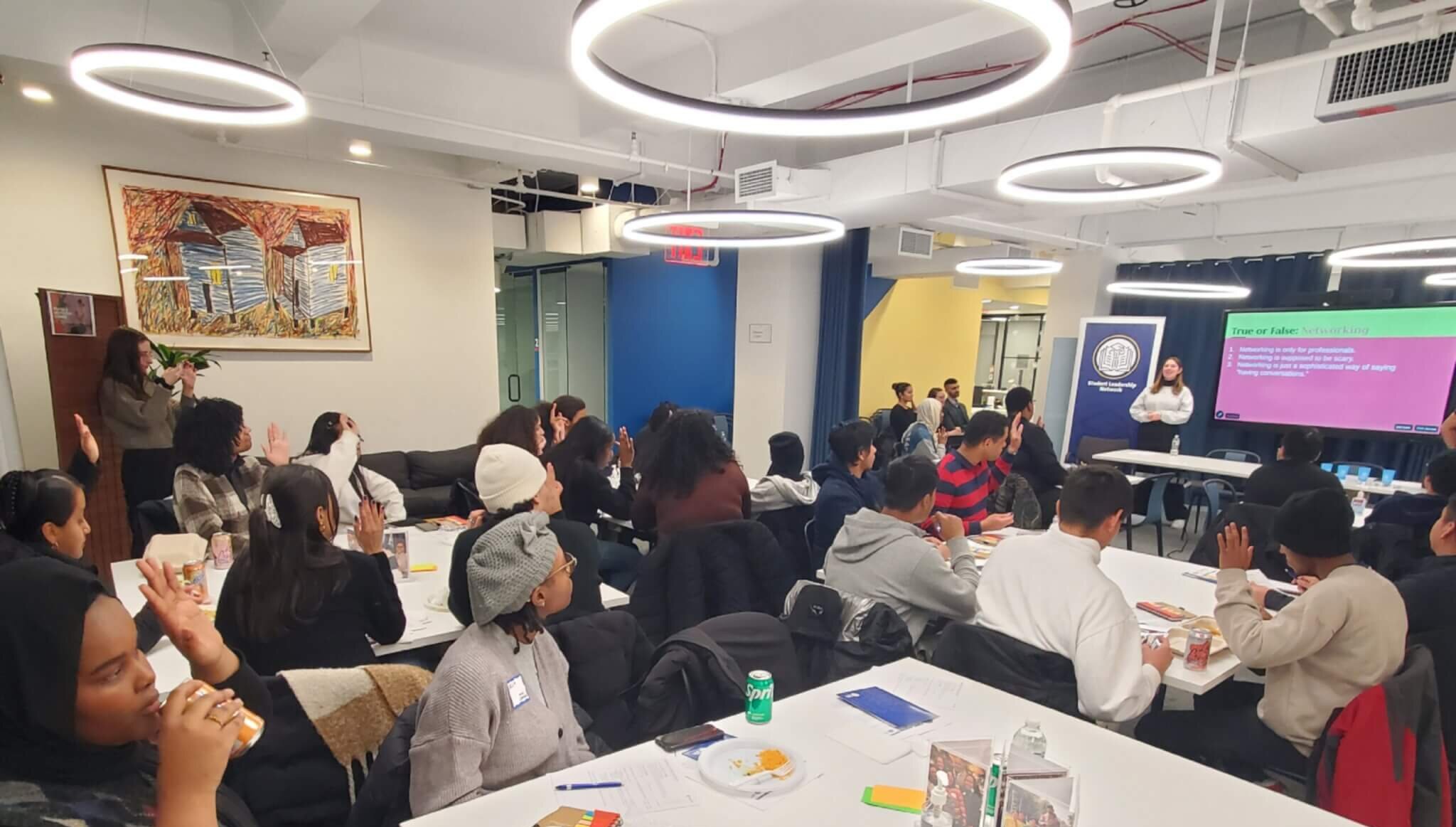 Students and alumni seated at small tables raise their hands in response to a prompt by the presenter at the front of the room