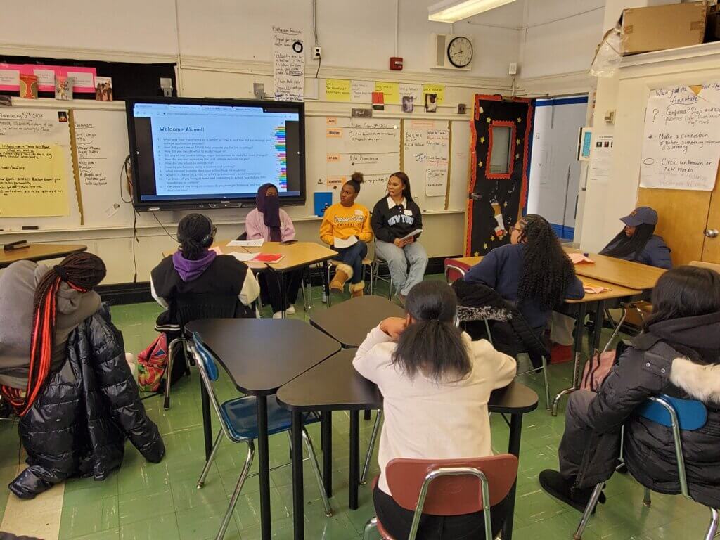 Three alumni sit in front of classroom, some wear college sweatshirts and one wears a hijab.