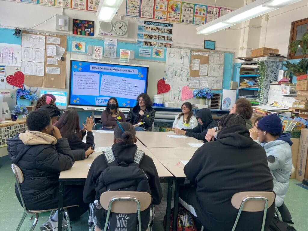 Alumni, teacher, and students are seated around a square table, with a screen behind them with title "Bronx Envision Academy Alumni Panel"