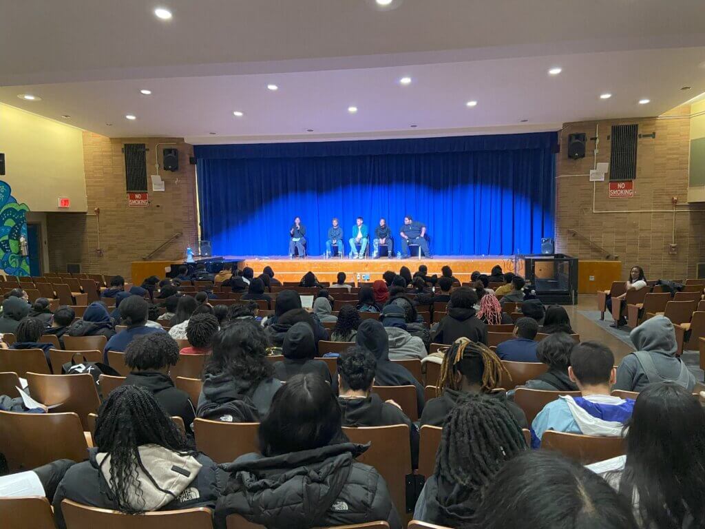 Alumni and college counselor are seated on stage, with a filled auditorium of seated students