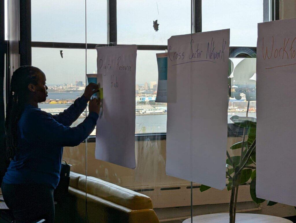Woman in a blue sweater and jeans with braids in her hair, affixes a post-it on one of three large poster papers