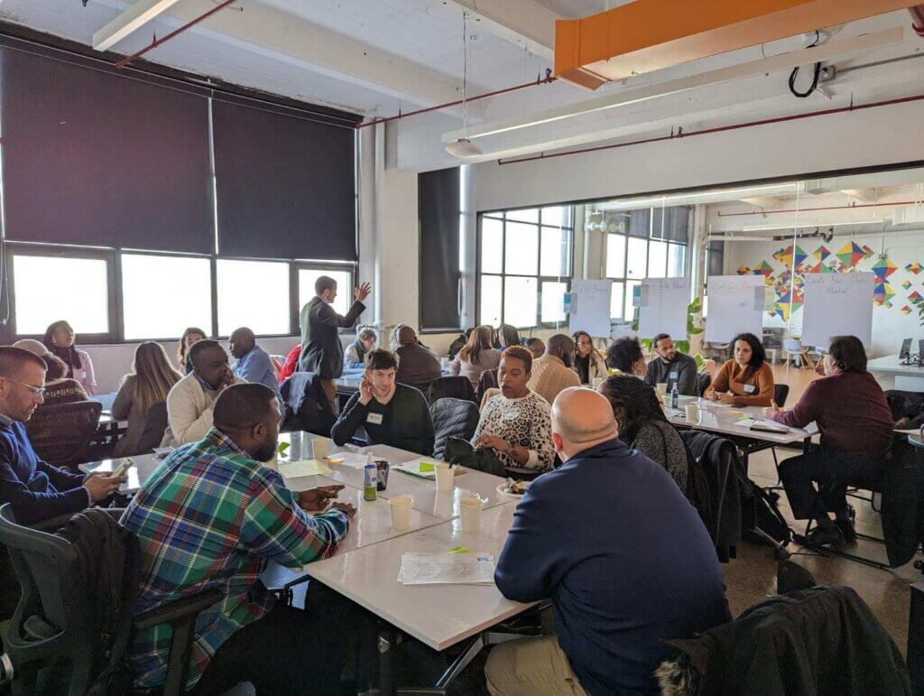 Adults are seated among four tables in a conference room, with one man standing and talking with a hand upraised