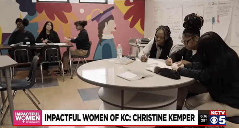 Groups of students sit at round tables in front of a colorful mural
