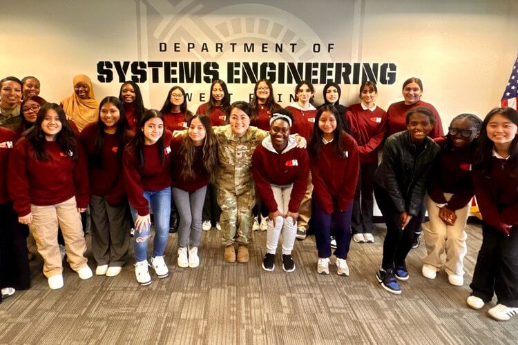 Students in uniform stand with uniformed staff at West Point Academy, in front of sign "Department of Systems Engineering"
