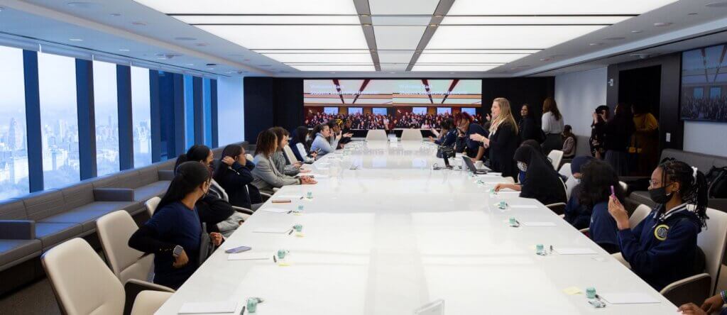 Students are seated around an oval boardroom desk, as Apollo staff member stands and talks