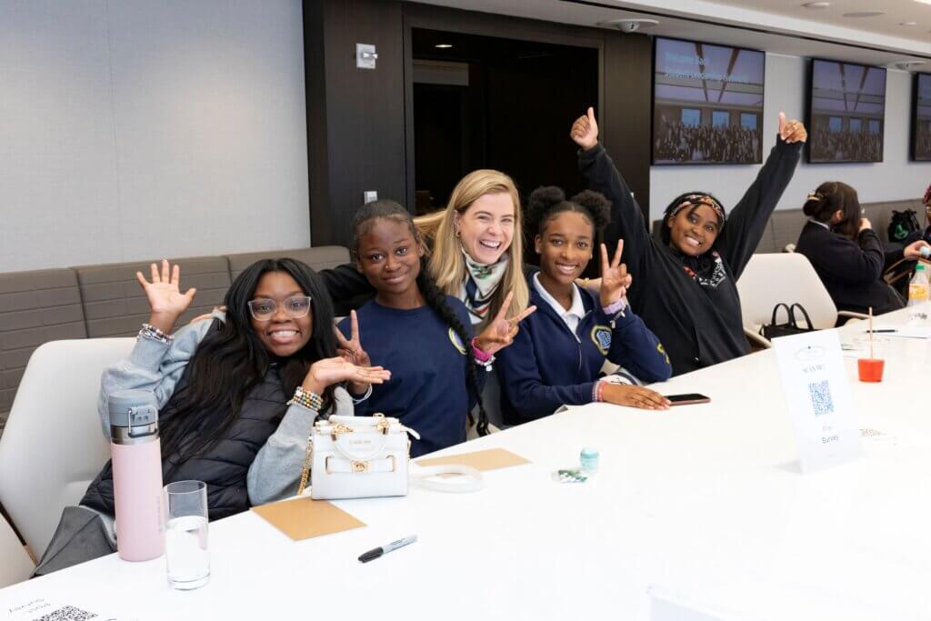 Four smiling students and an adult have thumbs up or peace signs with their fingers