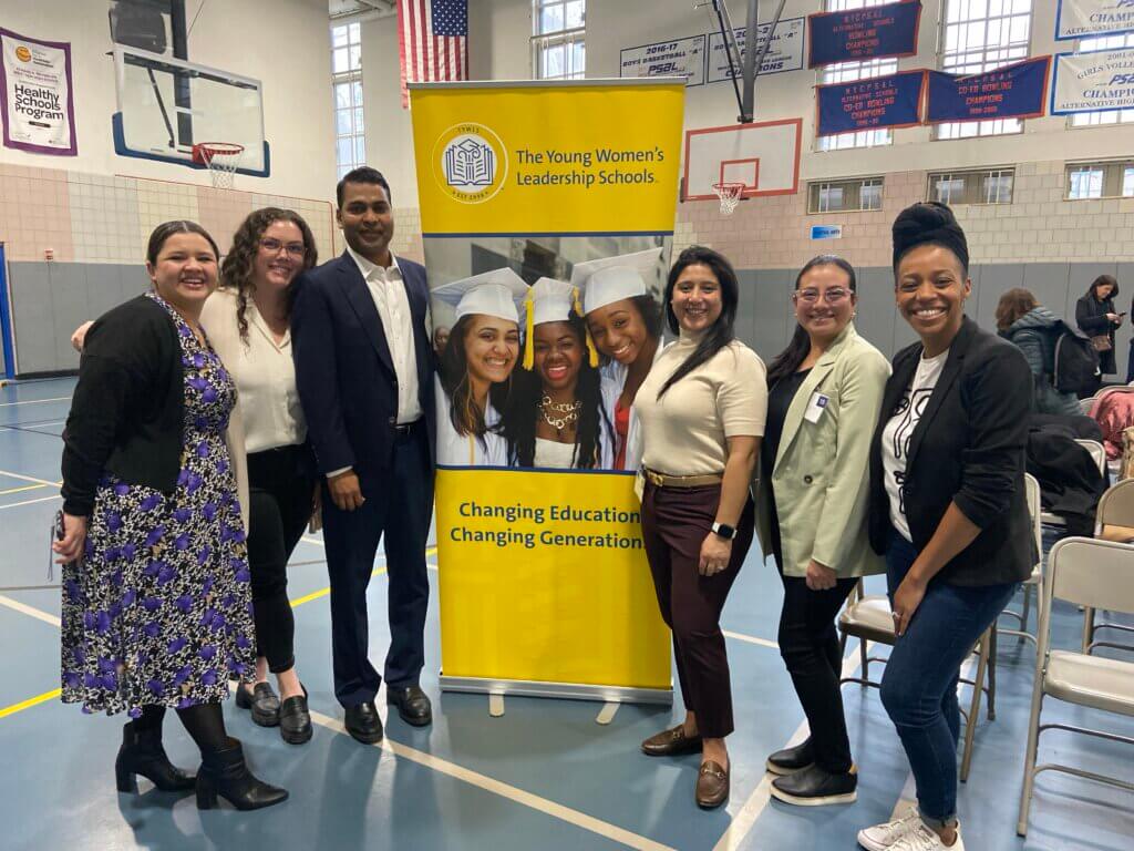 Six adult volunteers gather around a school sign in the gymnasium