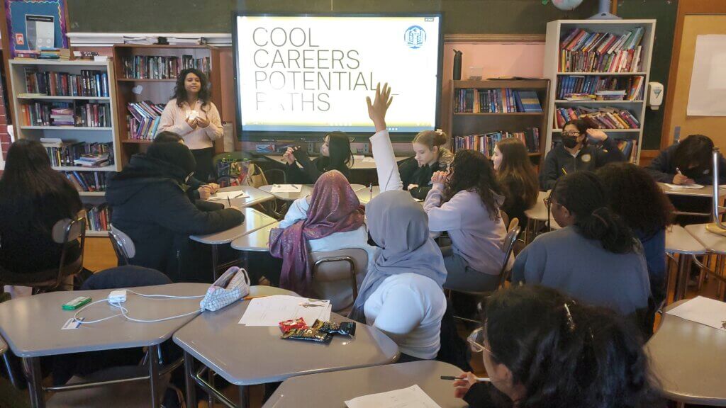 Student raises hand as volunteer looks to her from front of room, next to screen that says "Cool Careers Potential Paths"